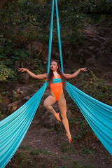 Beautiful aerialist girl doing acrobatic and flexible tricks on blue aerial silks tissues outdoors. Day light.