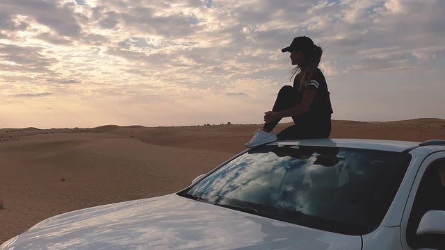 Young woman sitting on the roof of a car and looking on the epic sunset, against the background of the desert.