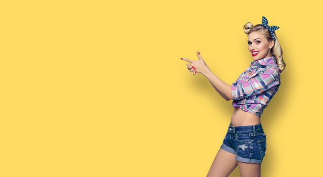 Happy young woman pointing. Excited girl in pin up style, showing something or copy space for advertise slogan or text. Retro fashion and vintage hairdo concept. Yellow color background.