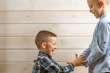 Fototapeta premium A 4-year-old boy in a blue klepy shirt cries on a light wooden background and his 10-year-old brother stands and plays rock-paper-scissors.