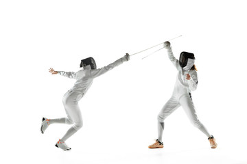 Fototapeta na wymiar Teen girls in fencing costumes with swords in hands isolated on white studio background. Young female models practicing and training in motion, action. Copyspace. Sport, youth, healthy lifestyle.