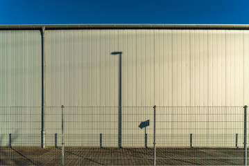 Fototapeta na wymiar Schatten von Straßenlaterne und Wegweiser auf einer Aluminiumfassade mit einem Zaun im Vordergrund