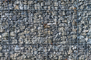 Gabion retaining wall. Metal wire grid filled of raw natural stones. Masonry exterior detail of protective barrier from metallic net cage with stacked gray rocks. Modern landscaping and architecture.