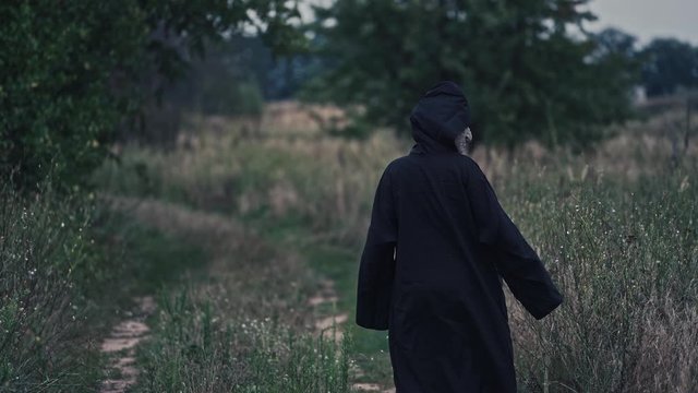 Backside portrait of death in nature. Dark figure in black costume out stretches its arms and goes away in the field in the evening. Scary nightmare ghost outdoors.