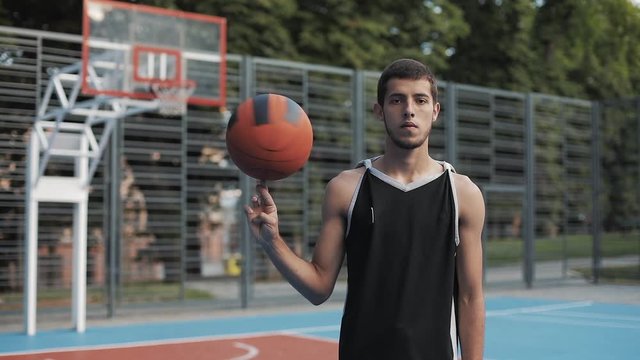 Young Caucasian Basketball Player Wearing Black Singlet Spinning the Ball on his Finger Standing at Street Basketbal Court and Looking to Camera. Healthy Lifestyle and Sport Concept.