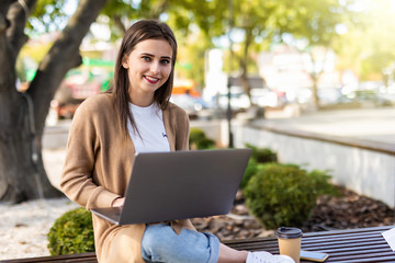 Beautiful young woman wearing autumn coat using laptop while sitting on a bench, drinking takeaway coffee cup