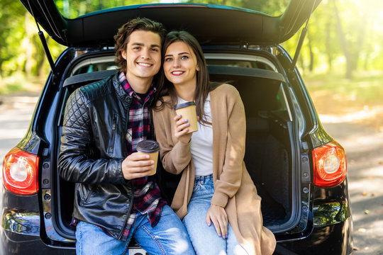 travel, summer vacation, road trip, leisure and people concept. Happy couple drinking coffee from disposable cups sitting on trunk of hatchback car outdoors