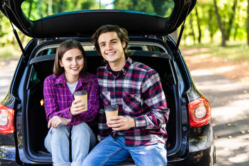 Beautiful young couple sitting with coffee in car trunk