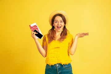 Happy holding tickets for traveling. Caucasian woman's portrait on yellow studio background. Beautiful model in cap. Concept of human emotions, facial expression, sales, ad. Summertime, travel, resort
