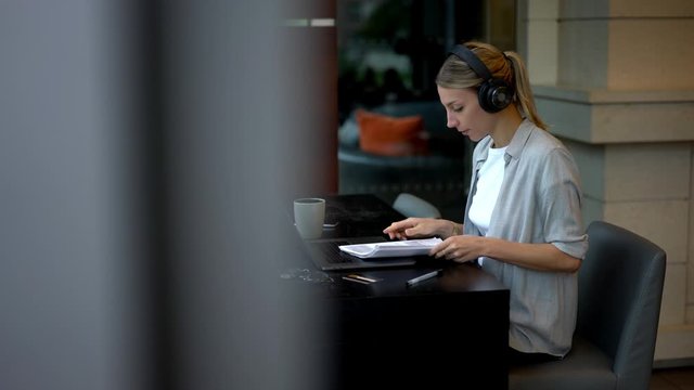 Intelligent female entrepreneur in electronic headphones for noise cancelation checking information from paper reports while sitting at desktop with laptop device and updating software