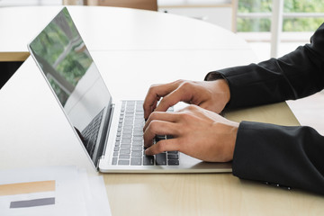 Close up on hand wearing black suit typing a laptop on a desk in an office.	