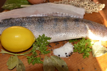 On a wooden board lies fresh pike perch fish fillet, yellow lemon, onion, salt, herbs, spices.