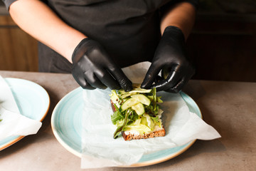 Obraz na płótnie Canvas Cook hands preparing and making sandwich. Preparing healthy vegetarian bruschettas in dark gloves. Sandwich with soft cheese, arugula and cucumber