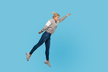 Fototapeta na wymiar Superman. Full length portrait of determined woman in sweatshirt and jeans flying up in air, feeling superpower and freedom, trampoline to success. indoor studio shot isolated on blue background