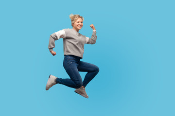 Obraz premium Full length portrait of woman with short hair in sweatshirt and jeans jumping or running in air, looking energetic and joyful, rushing for weekends. indoor studio shot isolated on blue background