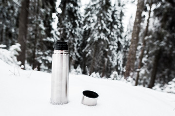 vacuum flask with hot drink on hill covered with snow near pine forest