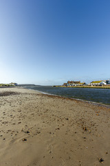 A view of a water channel between Christchurch (UK) harbor and bay with sandy beach and fishering buildings along the banks under a majestic blue sky