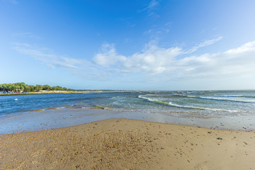 A view of a choppy water channel between Christchurch (UK) harbor and bay with sandy beach and trees along the bank under a majestic blue sky and some white clouds