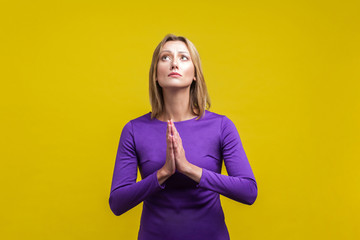 Please! Portrait of young woman in elegant purple dress holding hands in prayer and pleading, looking up with eyes full of hope, asking apology. indoor studio shot isolated on yellow background