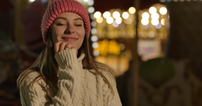 Charming Caucasian girl in winter hat and sweater standing in front of colorful carousels and looking at camera. Young woman spending weekends on city market. Cinema 4k ProRes HQ.