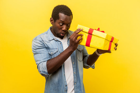 Long-awaited Interesting Gift! Portrait Of Happy Man In Denim Casual Shirt Unpacking Present, Looking Inside Box With Curious Happy Expression. Indoor Studio Shot Isolated On Yellow Background