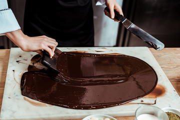 cropped view of chocolatier holding cake scrapers near melted dark chocolate on marble surface