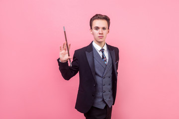 Portrait of young magician in tuxedo standing with raised magic wand and looking at camera with...