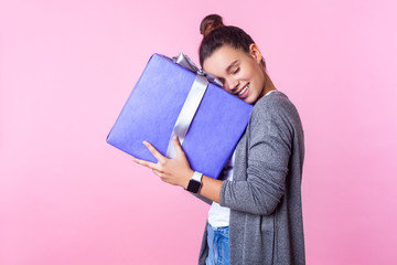 Long-awaited present. Portrait of delighted teenage brunette girl with bun hairstyle in casual clothes embracing gift box, satisfied pleased with surprise. studio shot isolated on pink background