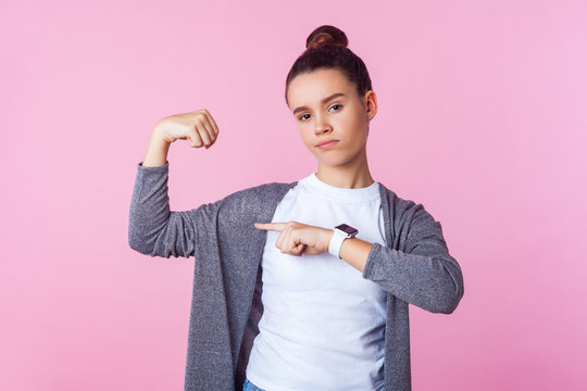Look at my strength! Portrait of confident brunette teenage girl with bun hairstyle in casual clothes pointing at her biceps, feeling powerful and independent. studio shot isolated on pink background