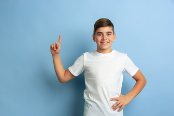 Smiling and pointing up. Caucasian boy portrait isolated on blue studio background. Beautiful teen male model in white shirt posing. Concept of human emotions, facial expression, sales, ad.