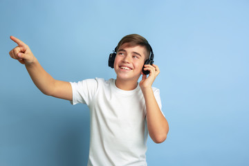 Listen to music, pointing at side. Caucasian boy portrait isolated on blue studio background. Beautiful teen male model in white shirt posing. Concept of human emotions, facial expression, sales, ad.