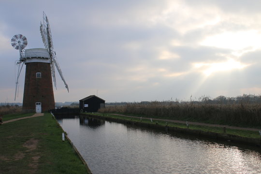 Beautiful Tranquil Landscape View Of Old Working Windmill Horsey Pump In Norfolk Broads East Anglia Uk People Stand By Still Waters Reflecting Fields Trees Red Brick With White Wood Sails In Grey Skie