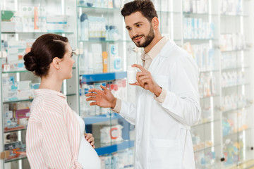 Smiling pharmacist showing to pregnant woman jar with pills by drugstore showcase