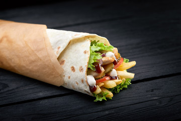 Shawarma rolled in lavash, moist grilled meat with onion, herbs and vegetables on wooden black background.