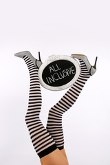 A female model's long legs are stretched out high in  a vertical form in front of a white background. Her  legs are decorated with a chalkboard sign with the words all inclusive written on it.