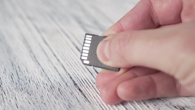 A young man holds a MICRO SD MEMORY CARD in his hand over a white textured wooden table and the light plays on electrical contacts
