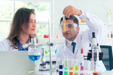 Male and Female scientist with a microscope in front of her reaching out for a test tube with blue solution in laboratory.