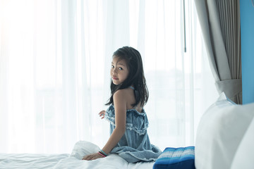 A bright, cute girl stood beside the bed in her bedroom. Beautiful little girl smiling. Portrait of cheerful kid lies at windowsill.