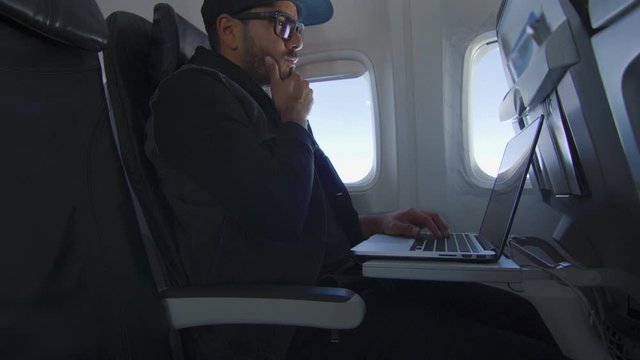 Man on airplane working on laptop, suddenly thinking of something great
