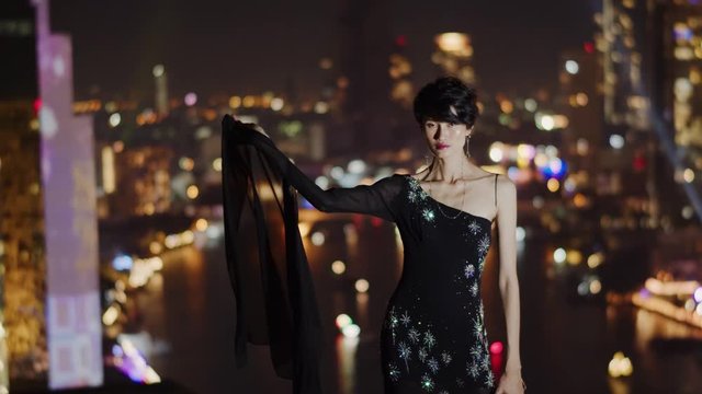 With the dramatic night lights and river in the blurred, bokeh background, a young Asian woman poses on a rooftop wearing a formal evening gown