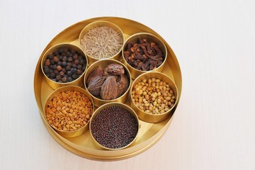 Spice box or collection of Indian spices or Garam Masala like cumin seeds, coriander, Fenugreek, big cardamom, black pepper, cloves, mustard seeds in designer brass container on wooden background