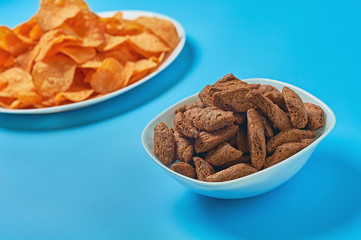 White ceramic plate and bowl full of snack for beer spicy potato chips, pieces of rusk bread lies on blue table in sport bar or pub. Close-up