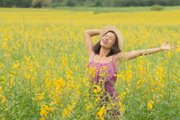 Naklejka na ściany i meble asia woman with a hat in her hand walks in a field with field flowers and smiles sincerely, happy enjoying summer in yellow field at sunset. smiling with arms raised up. concept of freedom.