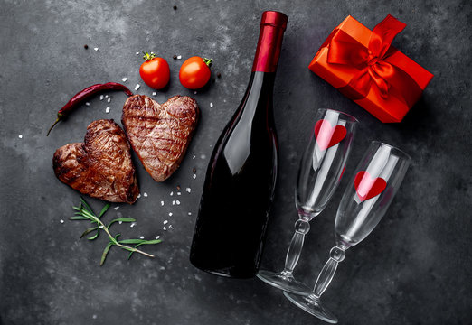two grilled beef steaks in the form of a heart with spices, a gift with a red ribbon and a bottle of champagne or wine with two glasses for dinner for Valentine's day on a stone background