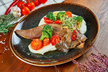 Close up on tasty Baked Beef Tongue Steak with Fresh Vegetables and Mashed Potatoes in black plate on wooden bakcground.