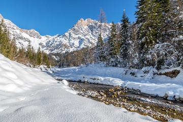 Fototapeta na wymiar Sunny winter landscape in the alps: Mountain range, river, snowy trees, sunshine and blue sky