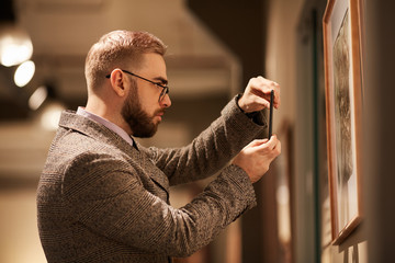 Side view of young bearded man in eyeglasses taking photo of paintings on his mobile phone in the gallery