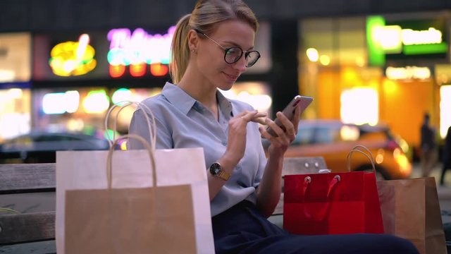 Smiling female buyer of purchases with Black Friday sales taking rest on city bench and searching next mall for visit and doing shopping, positive woman using cell smartphone for getting promo code