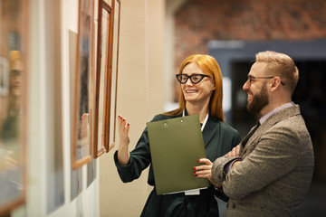 Art manager holding folder and holding the excursion for young man at art gallery they looking at pictures and discussing it - obrazy, fototapety, plakaty