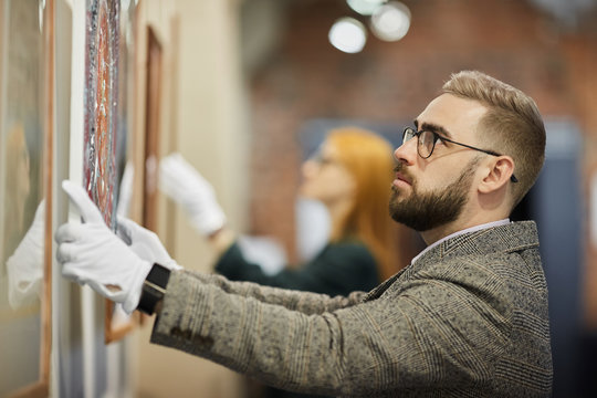 Bearded art manager in eyeglasses wearing gloves and hanging the painting on the wall with his colleague in the background